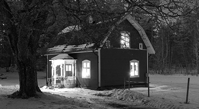 Maisonnette campagnarde un soir d’hiver, lumières allumées, avec arbre en avant-plan et forêt en arrière-plan (noir et blanc)