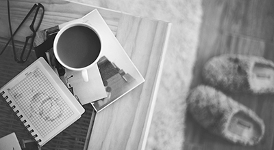 Une table avec tasse de café, carnet, photo et lunettes. Plancher avec pantoufles en arrière-plan (noir et blanc)