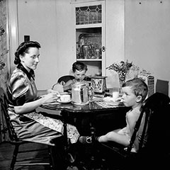 A mother and her sons are having breakfast. On the table, there is a toaster, a cereal box and a coffee maker.