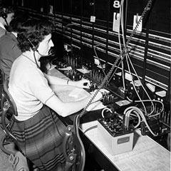 Une femme travaille comme téléphoniste devant une panneau de commande. Elle porte un casque d'écoute et un microphone.
