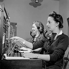Two women working as telephone operators. They wear a headset and a microphone allowing them to hear and speak. We can see in front of them the switchboard used for telephone communications.