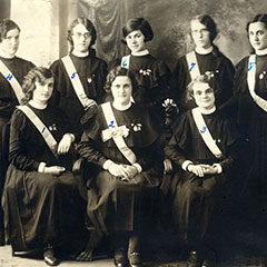 Photograph of a ribbon presentation ceremony. Seven young women pose proudly with their gowns and ribbons.