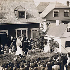 A parade float where a couple stands in front of a small house. Spectators are standing in the streets along the parade route.