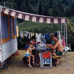 A family is eating on a picnic table under the shade of a folding camping trailer.