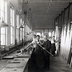 Un homme vêtu d'un habit explique à un ouvrier comment tailler un long tuyau. À l'arrière, deux ouvriers sont au travail.