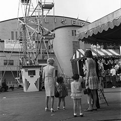 Deux mères et leur fillette sont devant la grande roue et un kiosque de jeux. À l'arrière, le Colisée de Québec est visible.