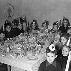 Children are gathered around a table decorated for a birthday party. They are wearing cardboard party hats.
