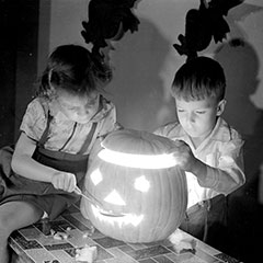 Two children are posing with a carved pumpkin. The girl is holding a spoon while the boy is putting the lid back on top.